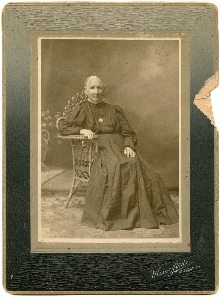 A monotone portrait of a seated aged woman with white hair and a mourning gown