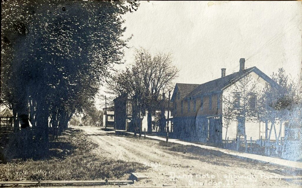 An old photo postcard showing a simple, two-story wood-sided structure on a dirt road. Other buildings, probably stores, are seen in the distance.